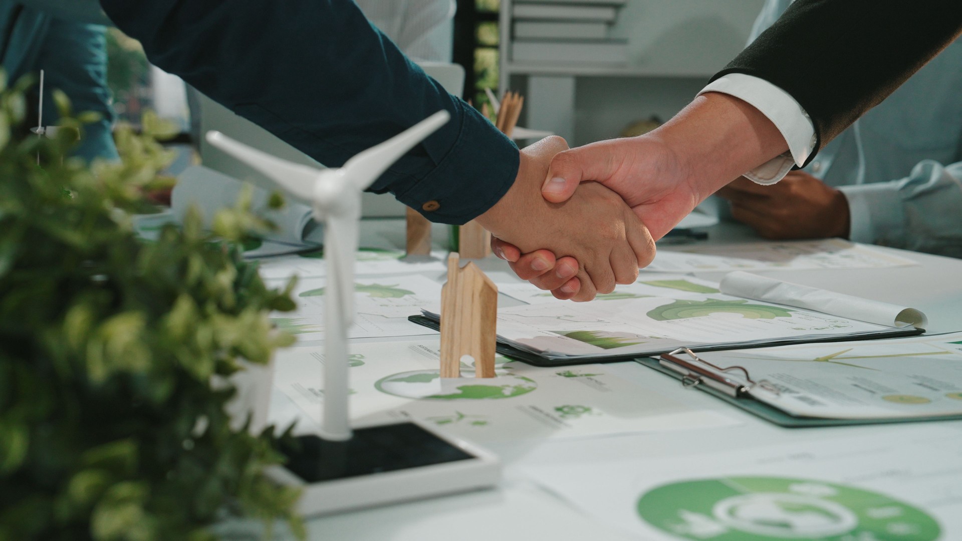 Hands joining in a business agreement with green energy models on the table, showcasing collaboration in sustainable development and corporate social responsibility SACTR