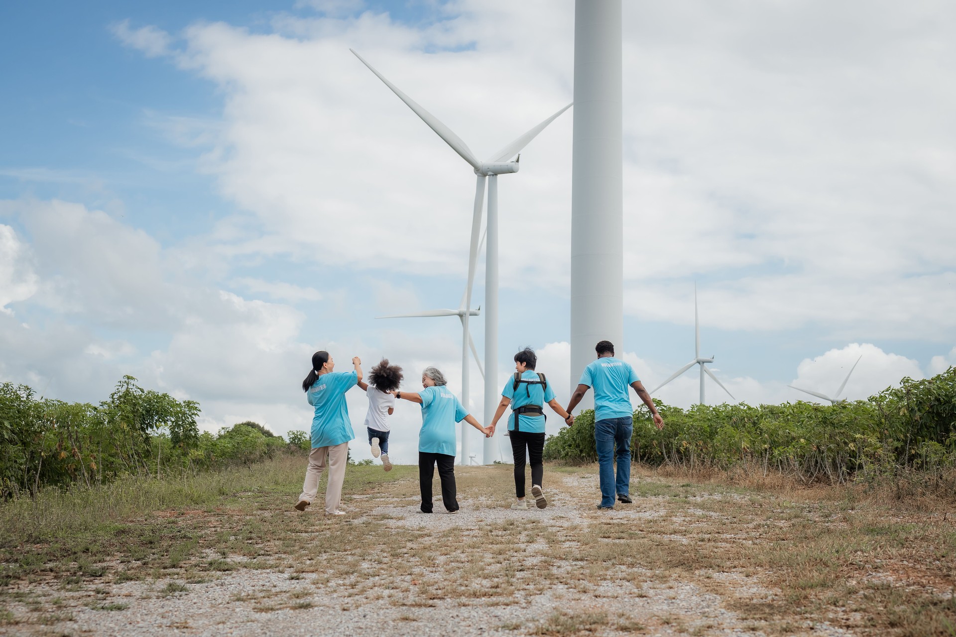 A group of people are walking together in a field, with a windmill in the background. Scene is calm and relaxed. environment, clean, wind turbine, volunteer, community, teamwork, social, care, nature